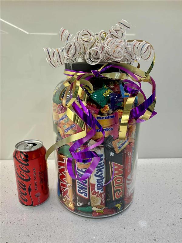 Large jar filled with assorted lollies wrapped with ribbon next to a soft drink can for scale
